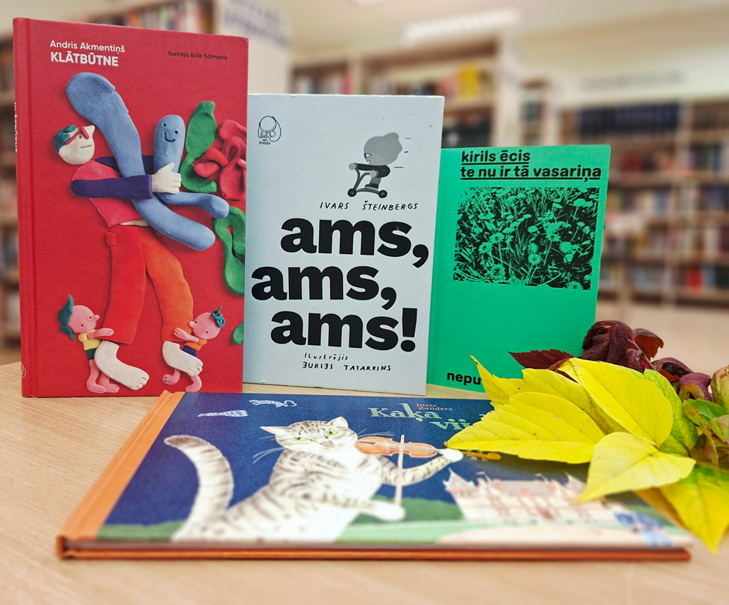 Četras grāmatas uz galda bibliotēkā, tostarp A. Akmentiņa "Klātbūtne", I. Šteinberga "Ams, ams, ams!" un I. Zanderes "Kaķa vijole", dekorētas ar rudenīgām lapām