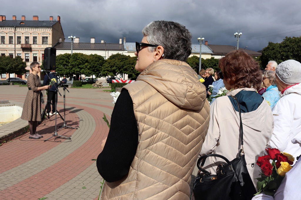 Skatītāji no mugurpuses vēro uzstāšanos pie mikrofona pilsētas skvērā