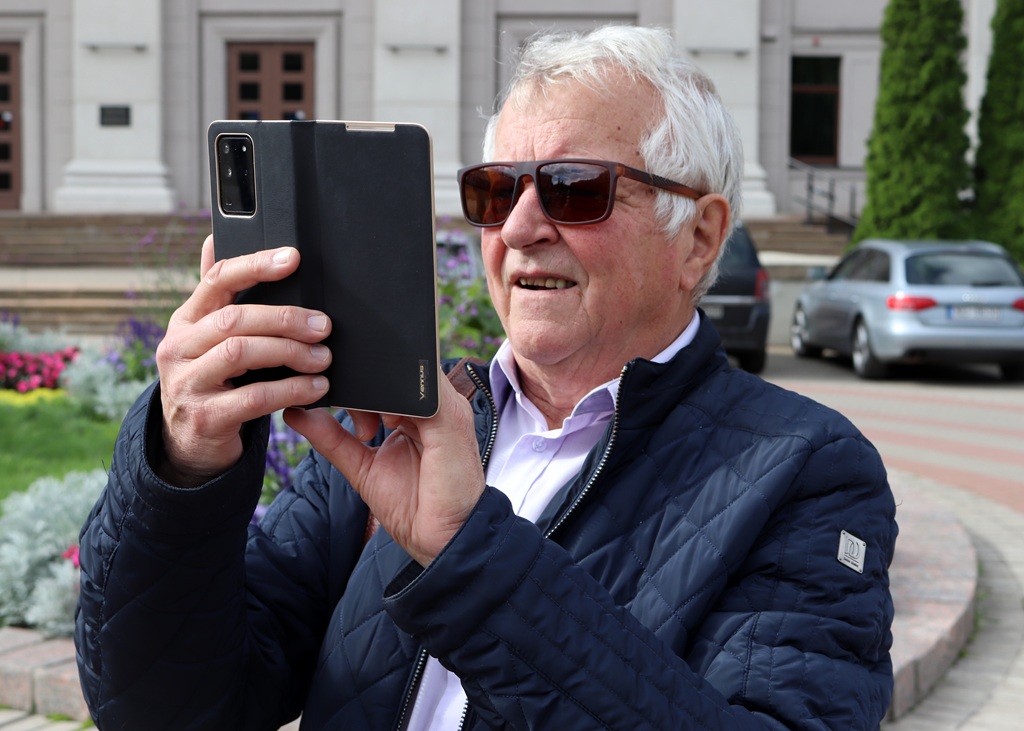 Senioru vecuma vīrietis ar saulesbrillēm fotografē ar viedtālruni ārtelpās pie ēkas