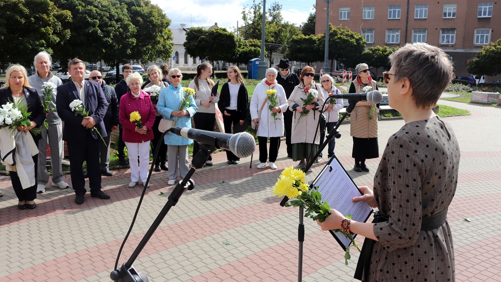 Sieviete uzstājas pie mikrofona brīvdabas pasākumā, uzrunājot klausītāju grupu ar ziediem rokās