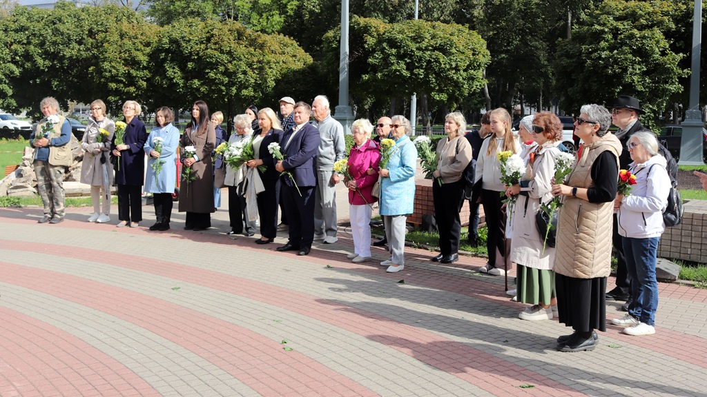 Plašs cilvēku pulks ar ziedu pušķiem rokās stāv puslokā brīvdabas pasākumā parkā