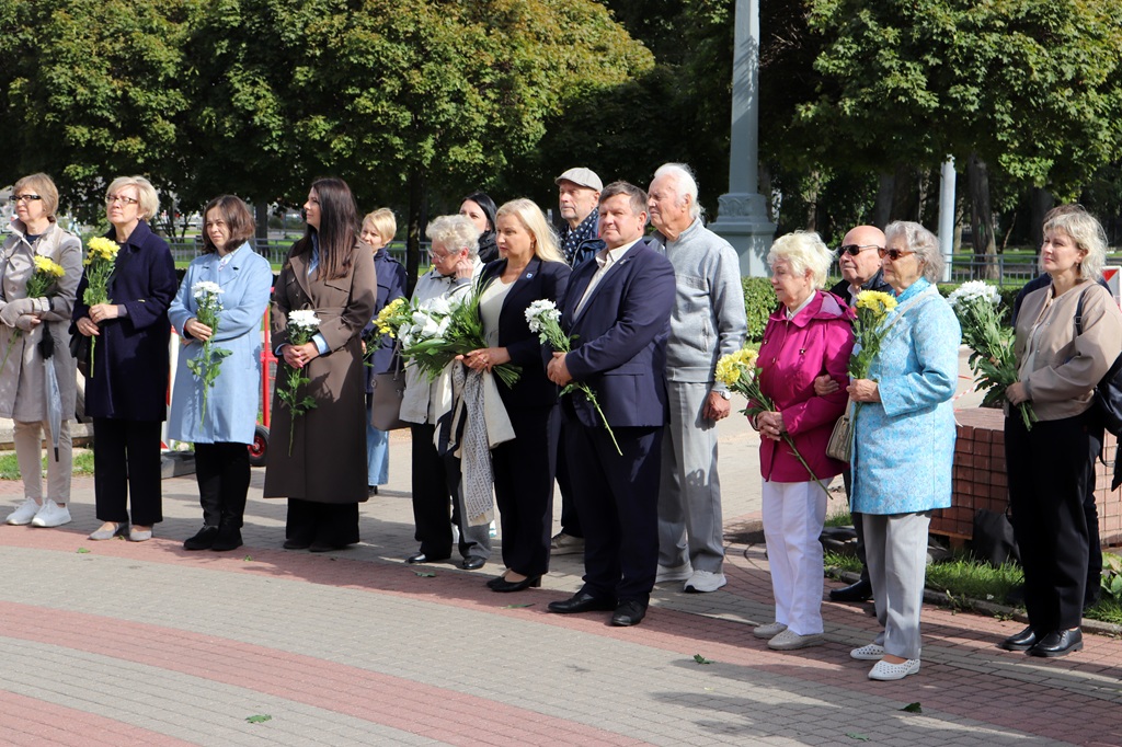 Pasākuma dalībnieku grupa ar ziediem rokās stāv rindā parkā ārtelpās