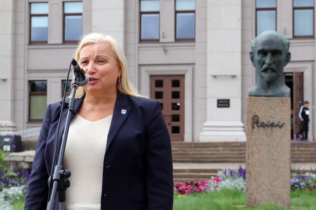 Sieviete tumši zilā žaketē uzstājas pie mikrofona brīvdabas pasākumā pie pieminekļa