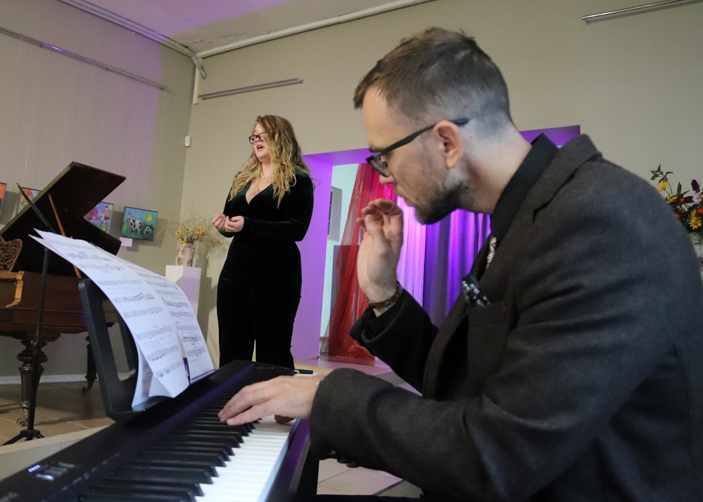 Pianists tuvplānā spēlē klavieres, kamēr sieviete izpilda dziesmu pasākuma zālē