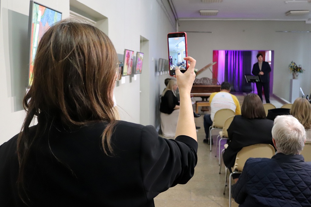 Sieviete no mugurpuses ar viedtālruni fotografē uzstāšanos bibliotēkas pasākumu zālē