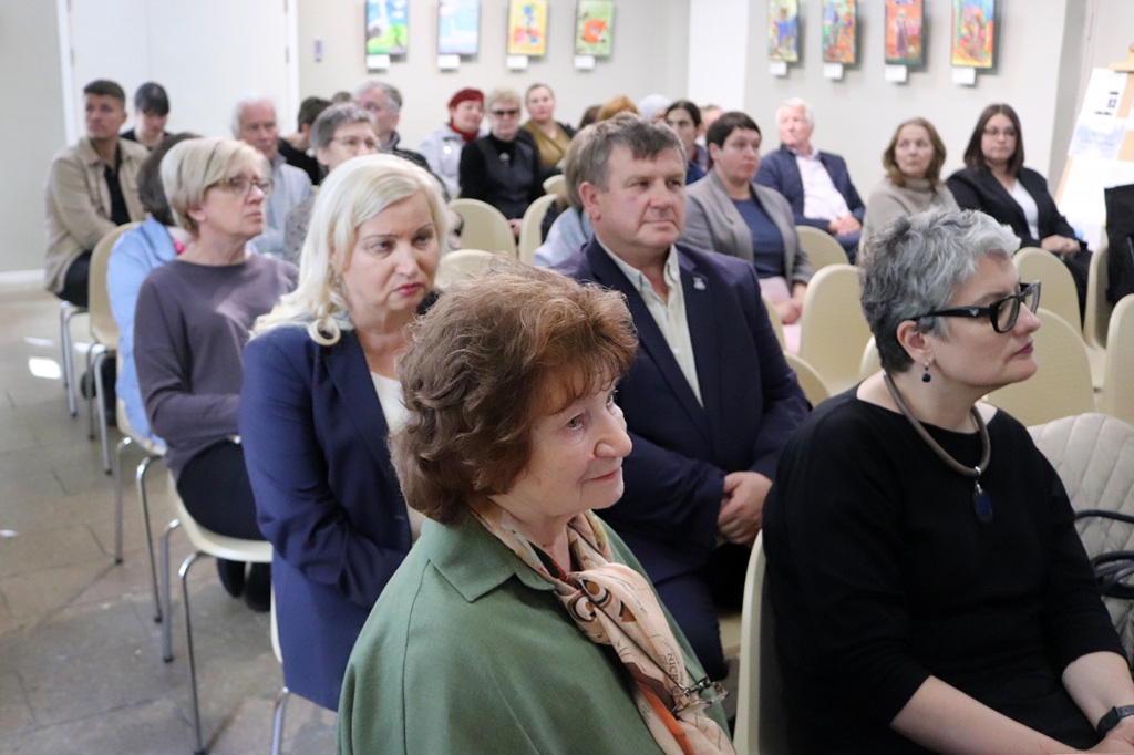 Skatītāji sēž krēslos un uzmanīgi vēro pasākuma norisi bibliotēkas zālē