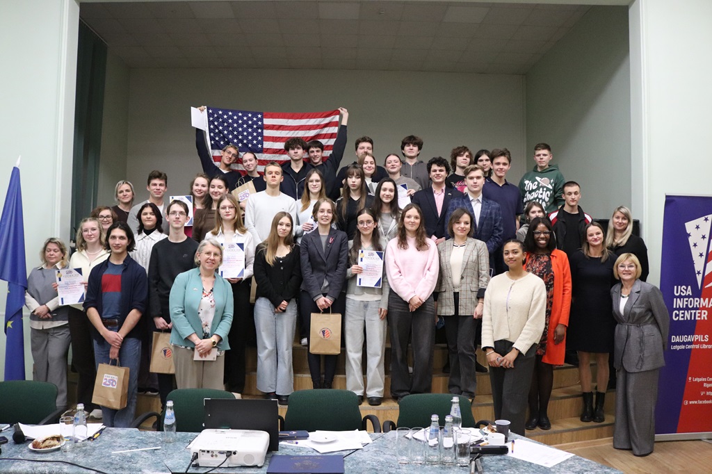 Liela dalībnieku un organizatoru grupa stāv un smaidīgi pozē fotogrāfijai telpā ar ASV karogu augšā un informatīvo stendu "USA Information Center" labajā pusē