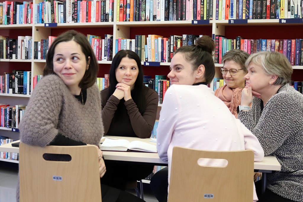 Sieviešu grupa sēž pie galda un uzmanīgi skatās uz priekšu bibliotēkas telpā ar grāmatu plauktiem fonā