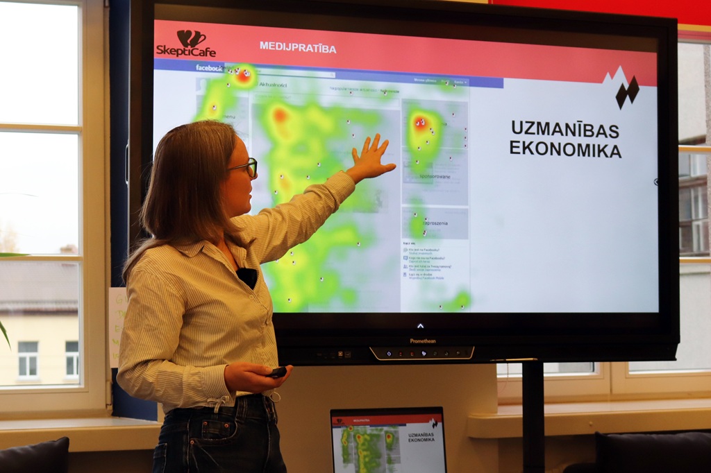 Sieviete ar brillēm un gaišā kreklā stāv pie interaktīvā ekrāna ar uzrakstu "UZMANĪBAS EKONOMIKA" un rāda ar roku uz attēla ar siltuma karti bibliotēkā