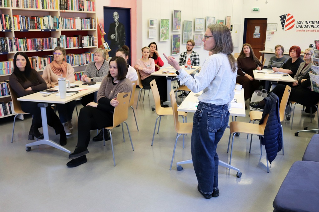 Sieviete gaišā kreklā un džinsos stāv zāles priekšā un runā ar dalībniekiem, kuri sēž pie galdiem bibliotēkas telpā ar grāmatu plauktiem un ASV Informācijas centra stendu