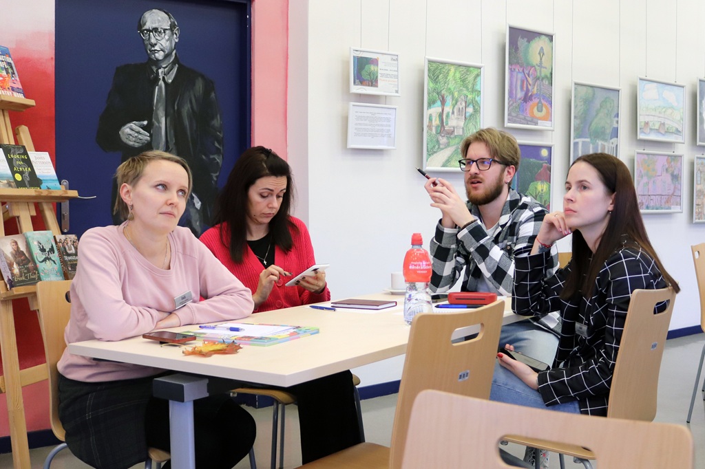 Četri semināra dalībnieki sēž pie galda un iesaistās nodarbībā, fonā redzami gleznojumi un bibliotēkas telpas interjers