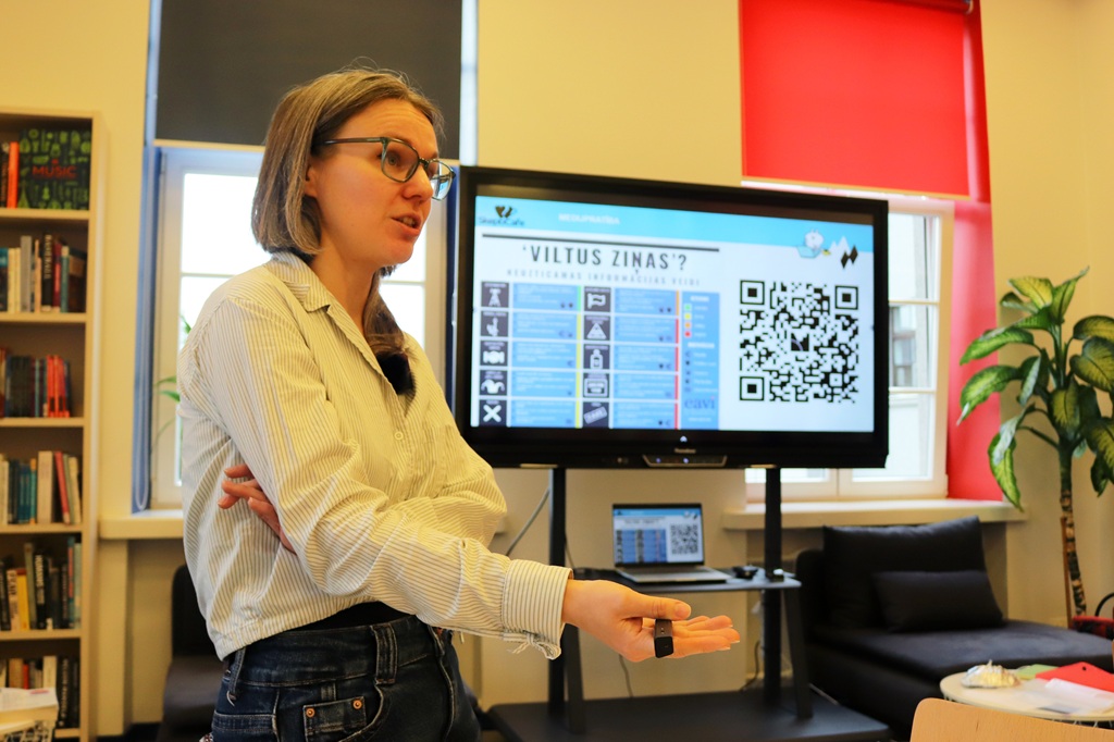 Sieviete ar brillēm un svītrotā kreklā stāv priekšplānā un runā, turot rokā pulti, kamēr fonā redzams ekrāns ar uzrakstu "'VILTUS ZIŅAS'?" un QR kodu