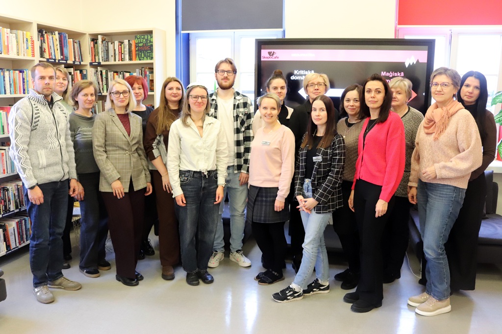 Liela semināra dalībnieku un lektori grupas fotogrāfija bibliotēkas telpā ar grāmatu plauktiem un ekrānu fonā