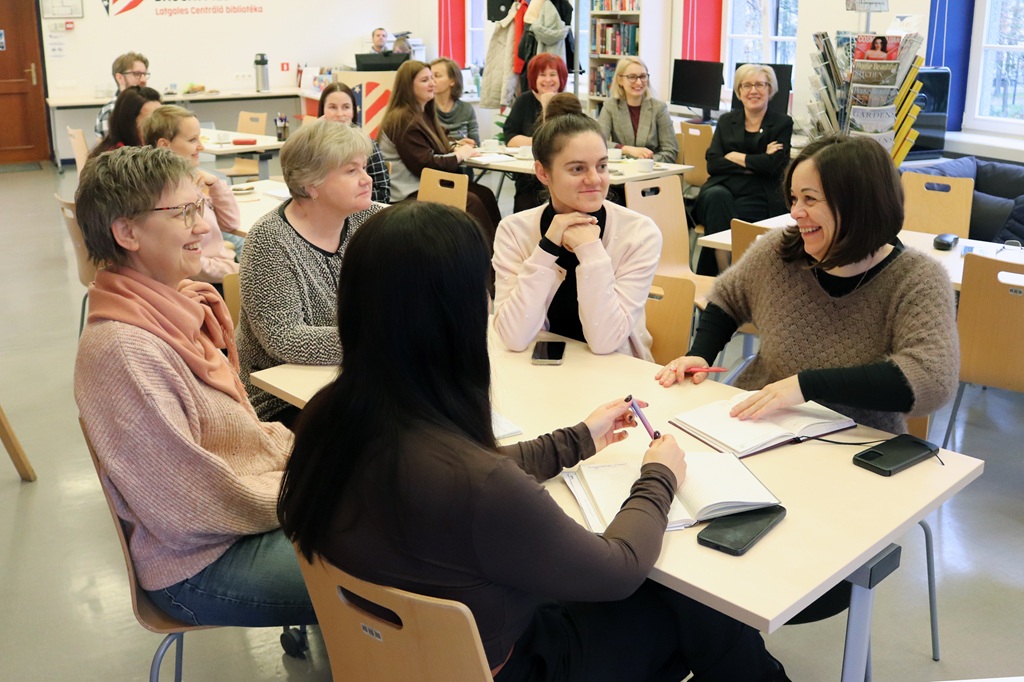Grupstiņa sieviešu sēž un smaidot apspriežas pie galda bibliotēkas telpā, kur fonā redzami citi apmeklētāji un plaukti