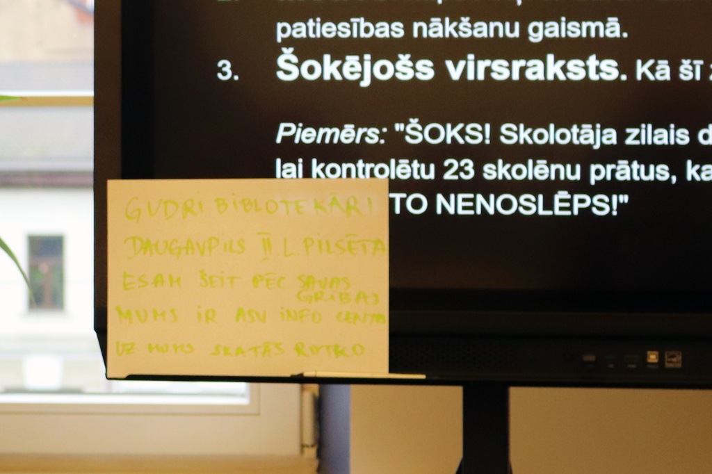 Ekrāns ar tekstu par šokējošiem virsrakstiem un uzlīme ar uzrakstiem "GUDRI BIBLIOTĒKĀ!" un "DAUGAVPILS II.L.PILSĒTA" pasākuma laikā