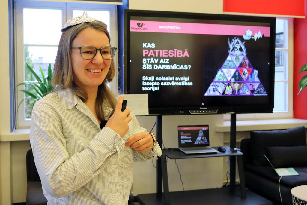 Sieviete ar brillēm un folijas cepurgalvu smaidot tur lapiņu un lasa tekstu ekrāna priekšā ar uzrakstu "KAS PATIESĪBĀ STĀV AIZ ŠĪS DARBNĪCAS?"