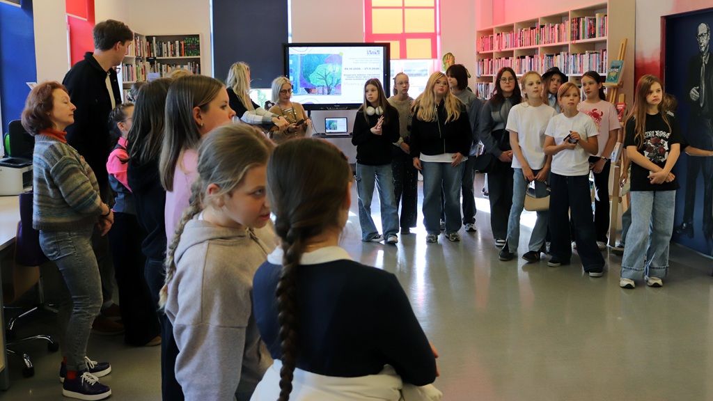 Liela bērnu un jauniešu grupa pulcējusies bibliotēkas telpā pie grāmatu plauktiem un ekrāna