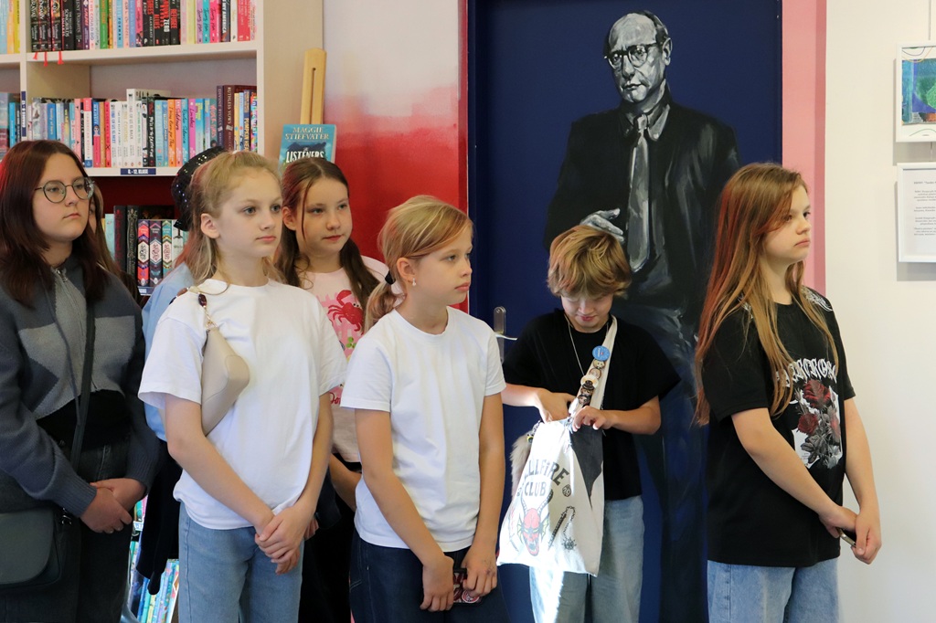 Skolēnu grupa stāv bibliotēkā pie grāmatu plaukta un apgleznotām durvīm