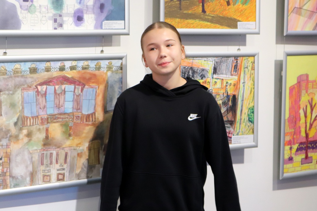 Jauna meitene melnā džemperī smaida, stāvot pie sienas ar krāsainiem zīmējumiem