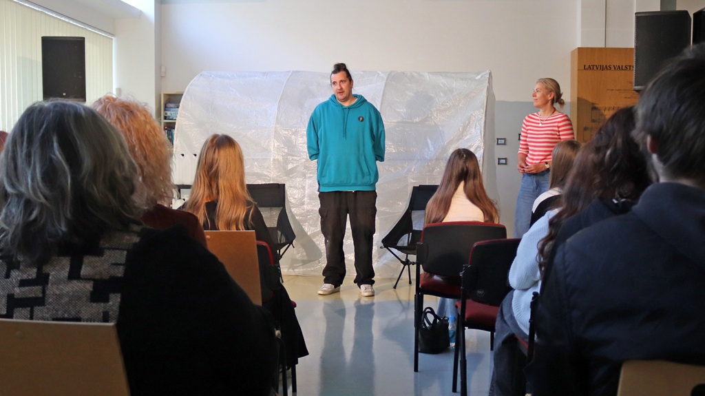 Vīrietis gaiši zilā džemperī un sieviete svītrotā kreklā stāv auditorijas priekšā gaišā telpā ar plastmasas plēves konstrukciju fonā