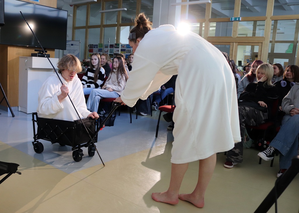 Jaunieši baltos halātos piedalās radošā performancē, kur viens no dalībniekiem sēž mazos ratiņos ar makšķeri rokās, kamēr otrs stāv blakus publikas priekšā