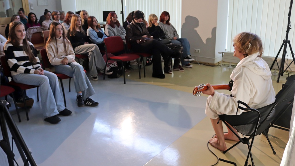 Jaunietis baltā halātā sēž krēslā ar ģitāru un uzstājas auditorijas priekšā, kamēr jaunieši sēž un uzmanīgi klausās pasākumu gaišajā telpā