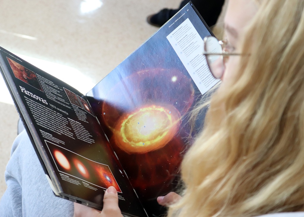 Gaišmataina meitene ar brillēm tur atvērtu grāmatu, kurā redzami kosmosa attēli