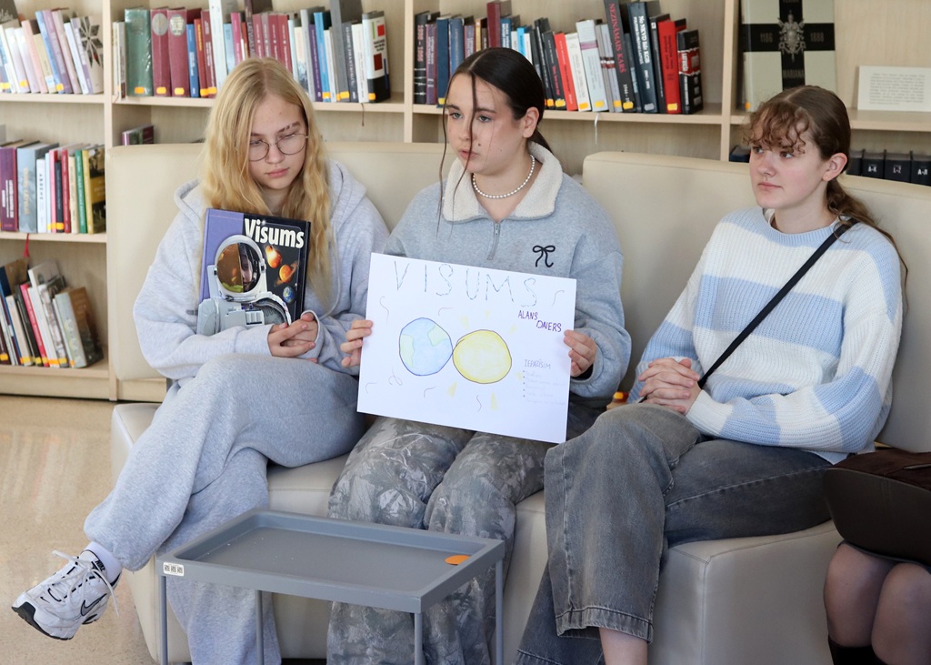 Trīs meitenes sēž uz dīvāna bibliotēkā, viena no viņām tur rokās plakātu