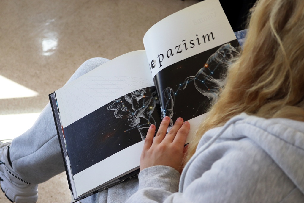 Sarmataina meitene pelēkā džemperī tur klēpī atvērtu ilustrētu grāmatu ar zvaigznājiem